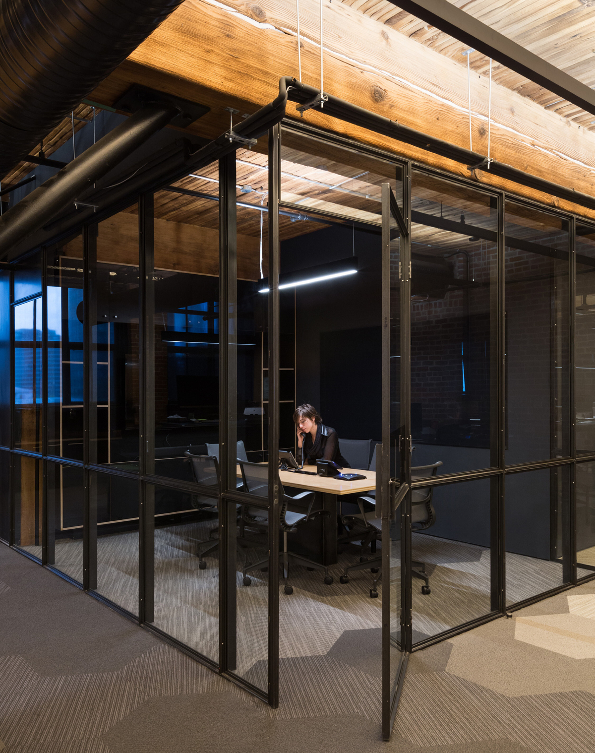 Interior evening view of Slack Headquarters, showing century-old timber post-and-beam construction with wood ceiling timbers above modern glass office and tech worker inside