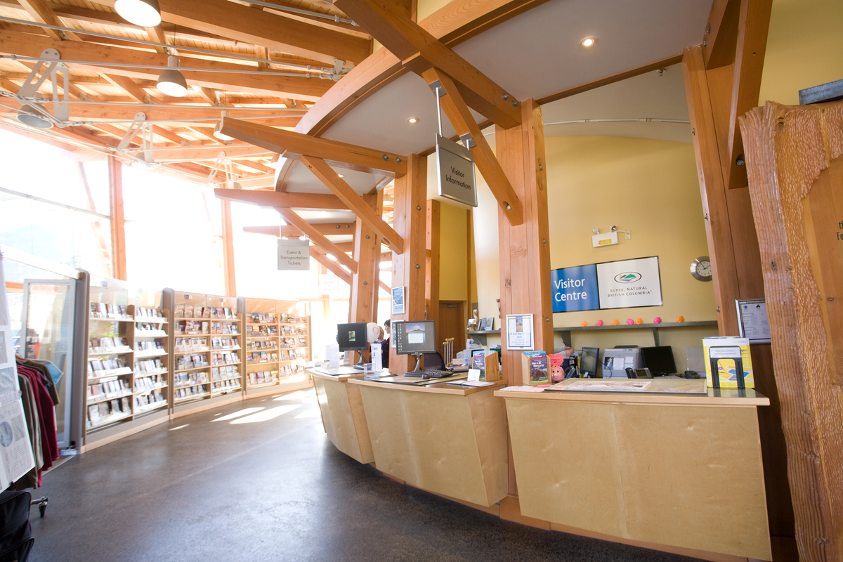 Interior view of Squamish Adventure Centre gift shop showing a roof of solid-sawn heavy timber beams supporting timber trusses and dimensional lumber roof slats; all above glazed outer wall and brightly lit B2C displays