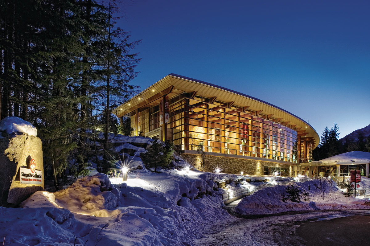 Exterior snowy nighttime view of low rise Squamish Lilwat Cultural Centre showing sweeping curved glass, Glue-laminated timber (Glulam), and post + beam timber elements
