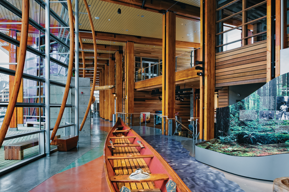 Interior daytime view of Squamish Lilwat Cultural Centre highlighting the wood floor, carved wooden canoe, and extensive use of mass timber in the columns, balcony, and ceiling structures