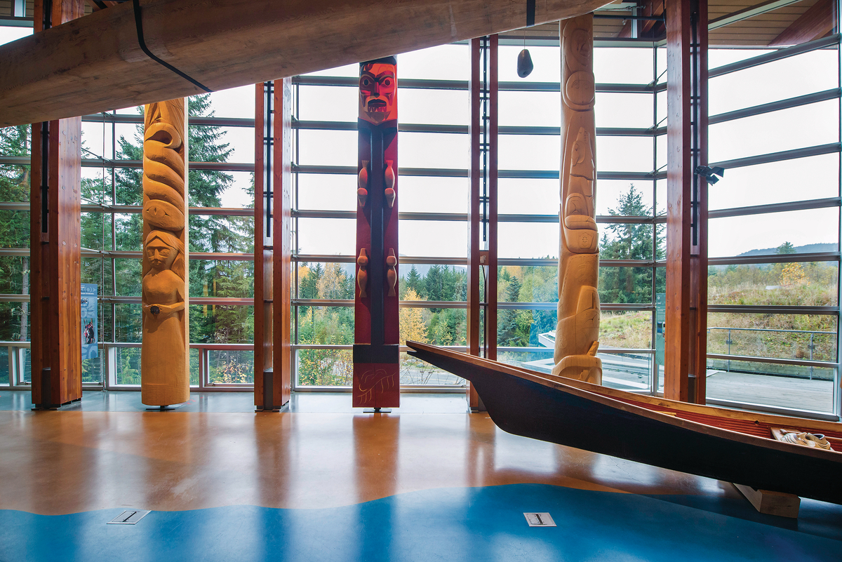Interior daytime view of Squamish Lilwat Cultural Centre highlighting wood floor, carved wooden canoe, and intricately carved First Nations vertical wood columns