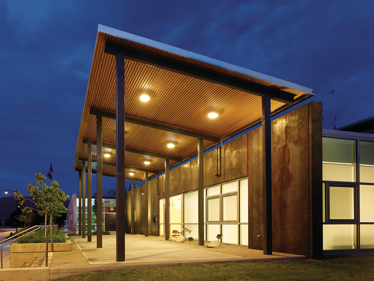 Overcast nighttime view of low rise Summerland RCMP Detachment showing exterior covered walkway with hybrid wood & metal construction featuring soffits clad with unfinished pine planks
