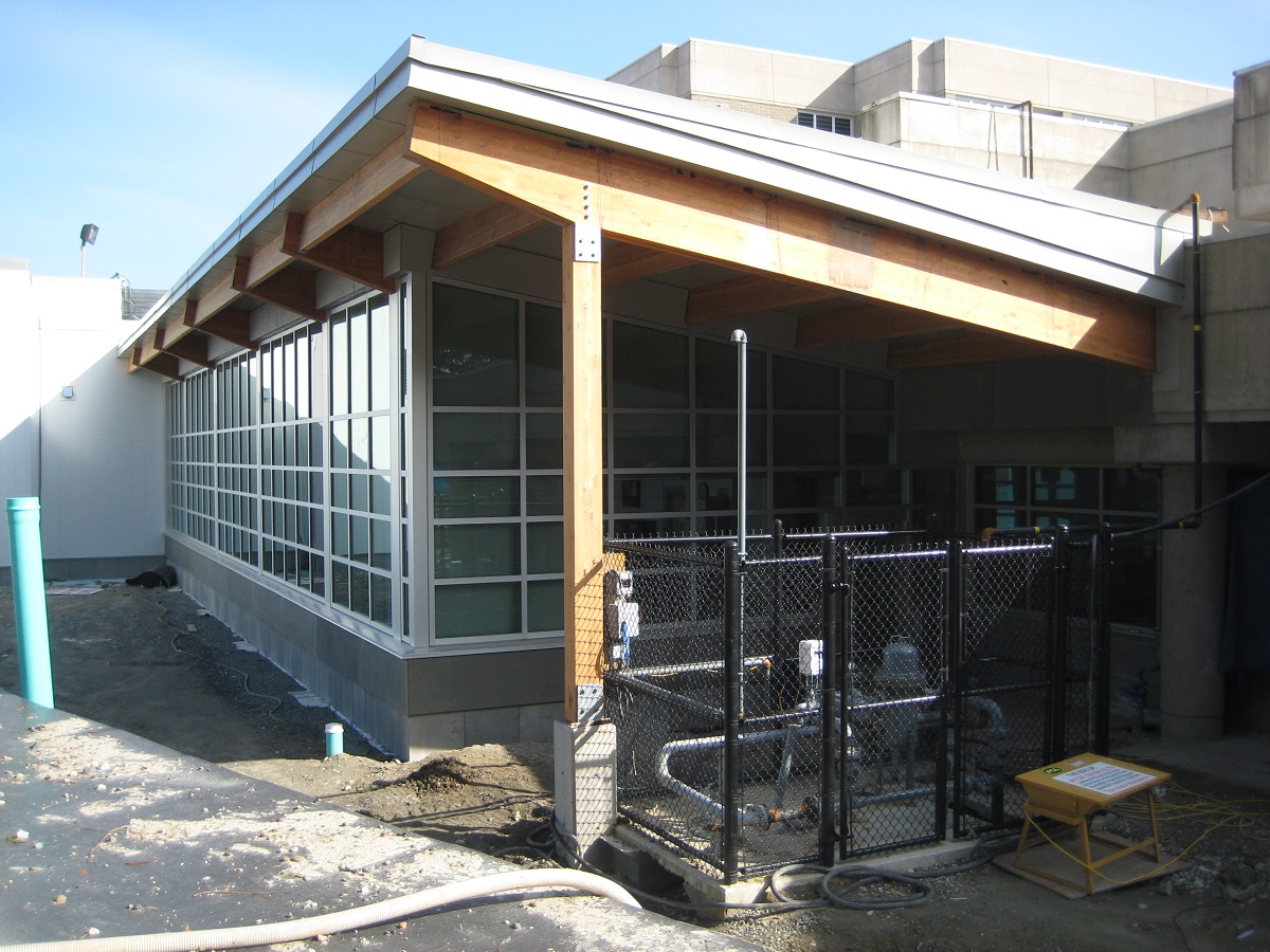 Exterior sunny late construction daytime view of close up of single story segment of Surrey Pretrial Services Centre showing metal, glass, and wood hybrid elements including a roof supported by Glue-laminated timber (Glulam) beams and columns