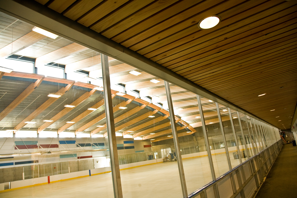 Interior daytime view of Trout Lake Ice Rink glassed in hallway with dimensional lumber slat ceiling looking into rink with glue-laminated timber (glulam) roof beams
