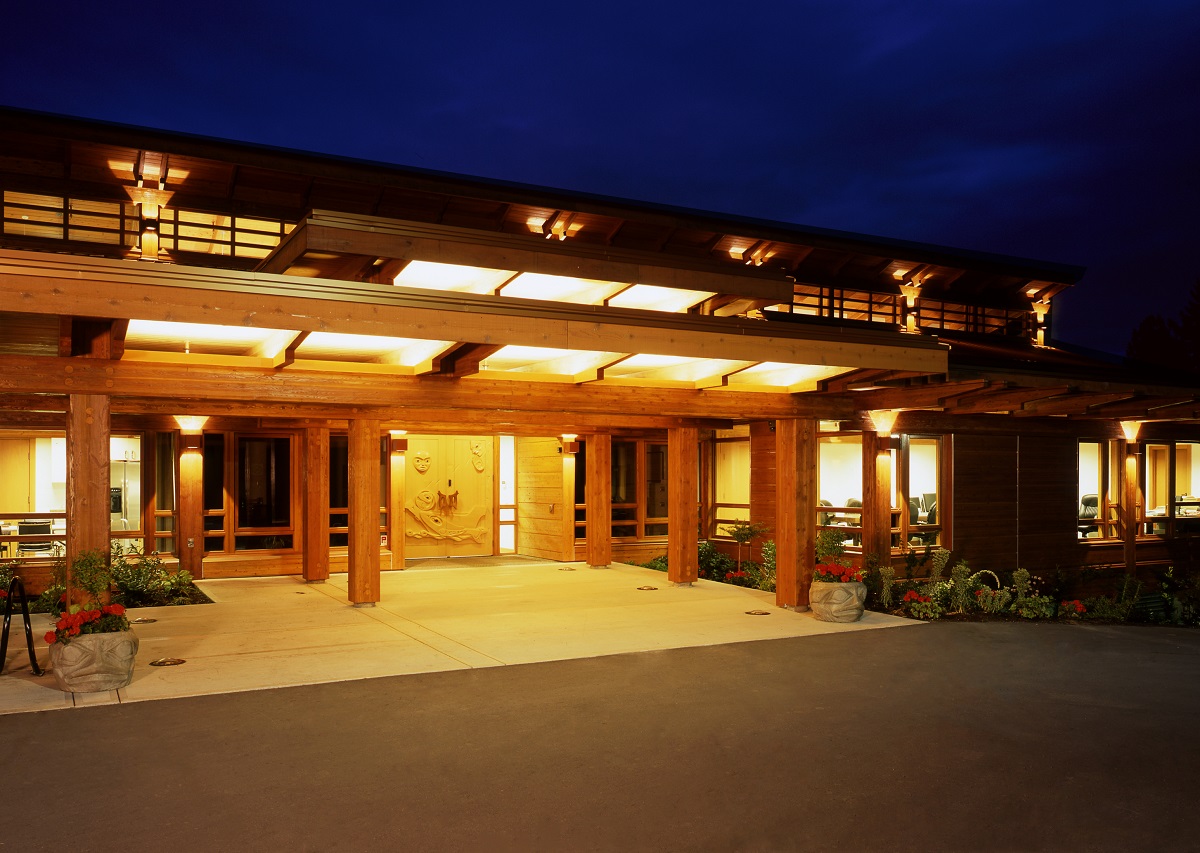 glue-laminated timber (glulam), millwork, paneling, and siding are all show in this nighttime exterior main entrance image of the Tseshaht Tribal Multiplex and Health Centre which highlights the abundant use of exterior wood for the column supports, hand carved doors, window trim, roof trusses, eves, and more