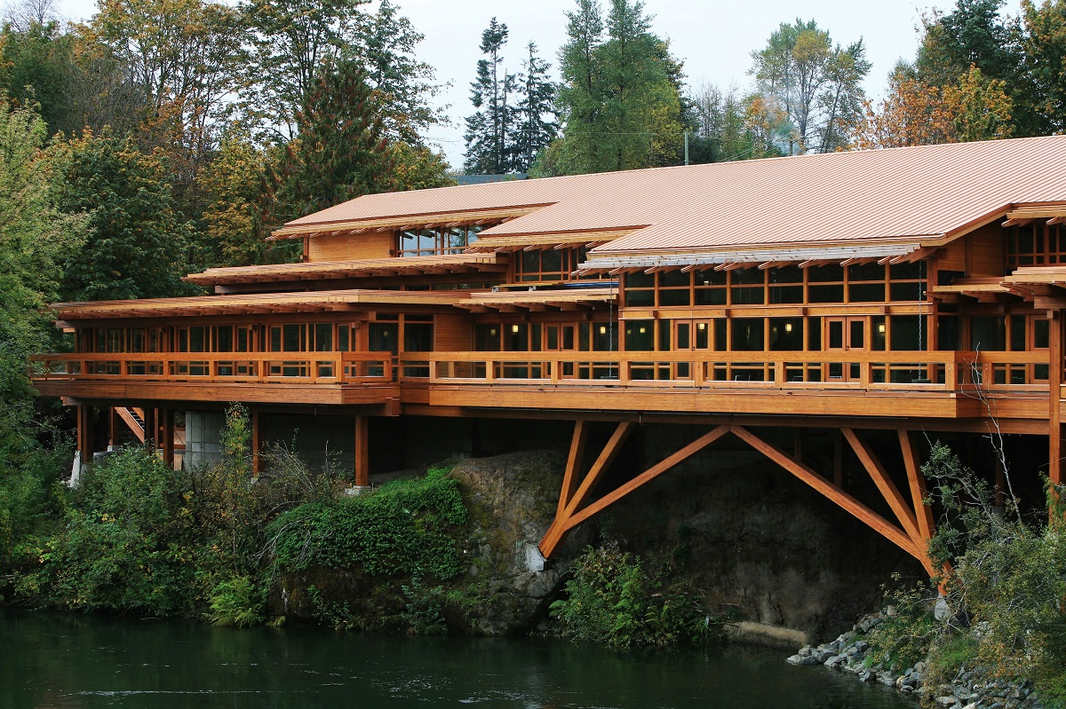 glue-laminated timber (glulam), millwork, paneling, and siding are all show in this cloudy exterior lakeside image of the Tseshaht Tribal Multiplex and Health Centre which highlights the abundant use of exterior wood for the column supports, wrap around balcony, window trim, roof trusses, eves, and more