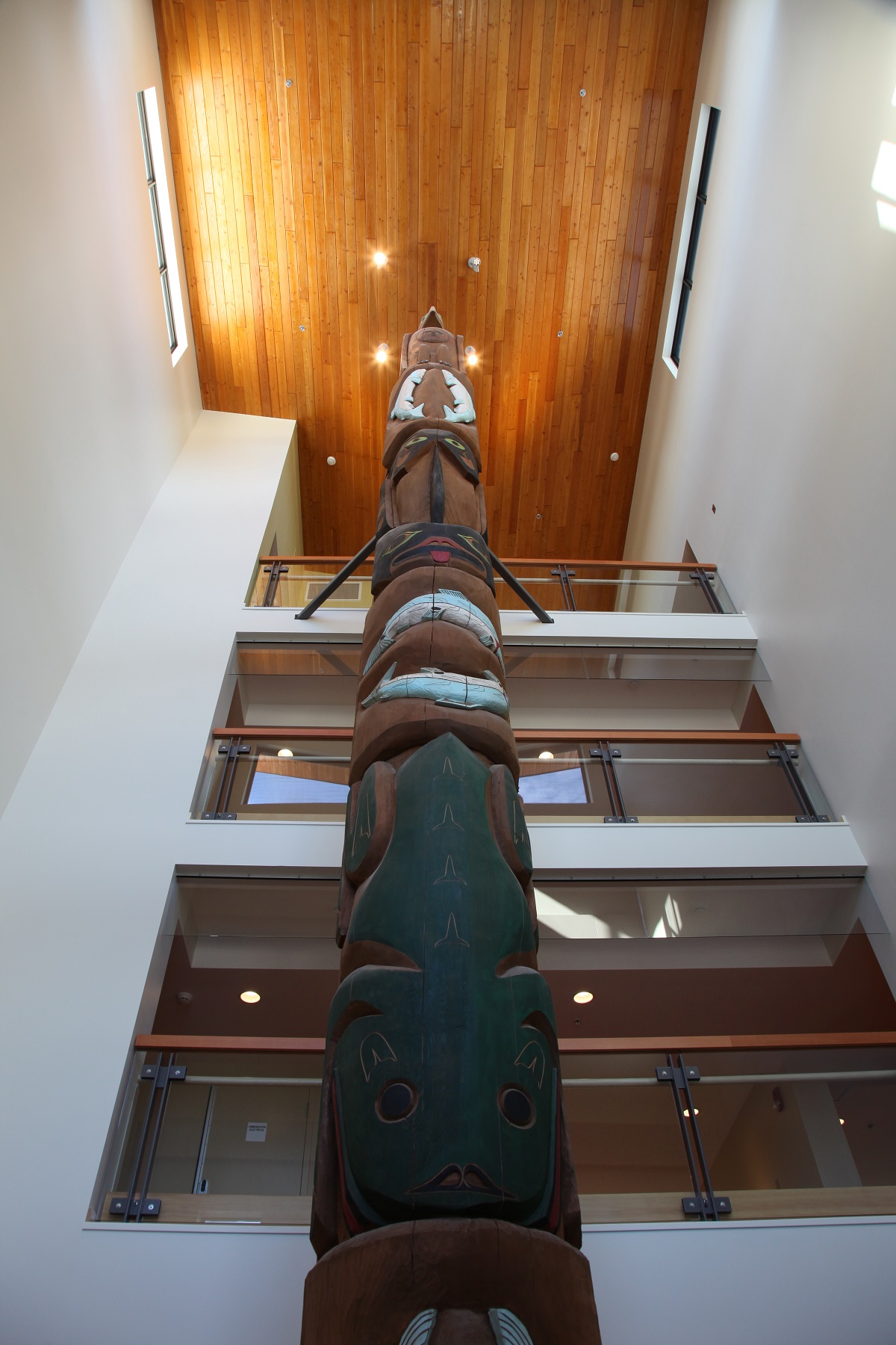 Interior upward daytime view of four-storey Ts'i'ts'uwatul' Lelum Assisted Living Facility main entrance atrium highlighting wood use for hand carved totem pole, balcony railings, and dimensional lumber ceiling