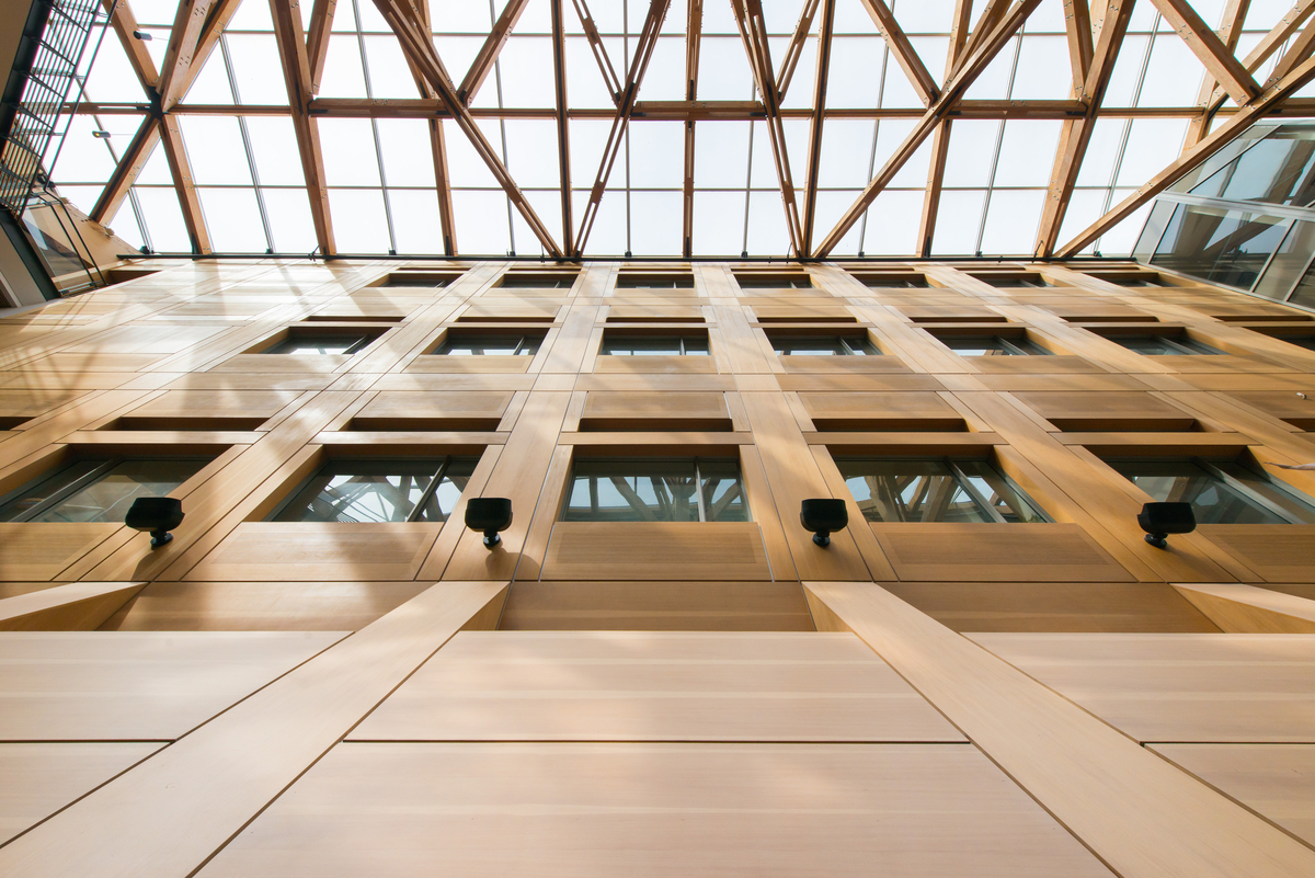 Interior daytime view looking up in multi-storey atrium of UBC Forest Sciences Centre with decorative wood paneling vertical surface, and intricate umbrella support trusswork of mass timber and glass roof above