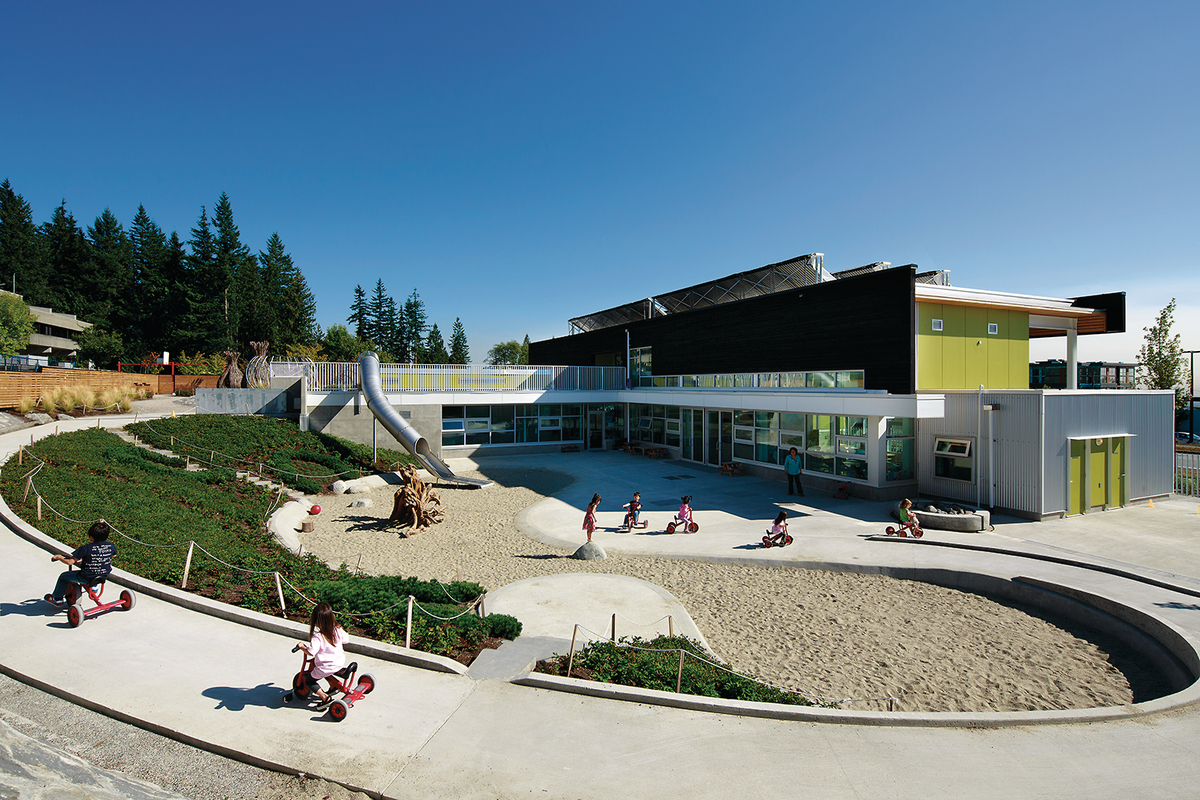Sunny daytime exterior photo of children playing outside UniverCity Childcare Centre, a net-zero energy building clad with glass, metal and western red cedar