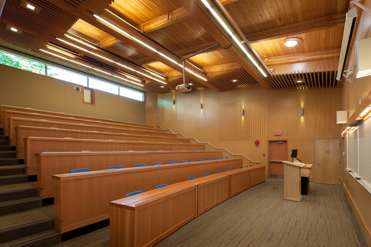 glue-laminated timber (Glulam) beams supporting tongue and groove ceiling lumber are all shown in this daytime interior image of a Vancouver Island University Cowichan Campus lecture hall, complete with blue plastic chairs at wooden counters configured as auditorium seating
