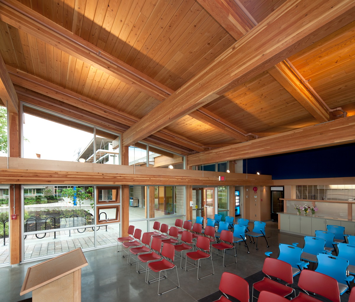 glue-laminated timber (Glulam) beams supporting tongue and groove roof lumber are all shown in this daytime interior image of the multi storey Vancouver Island University Cowichan Campus main assembly hall, complete with red and blue plastic chairs