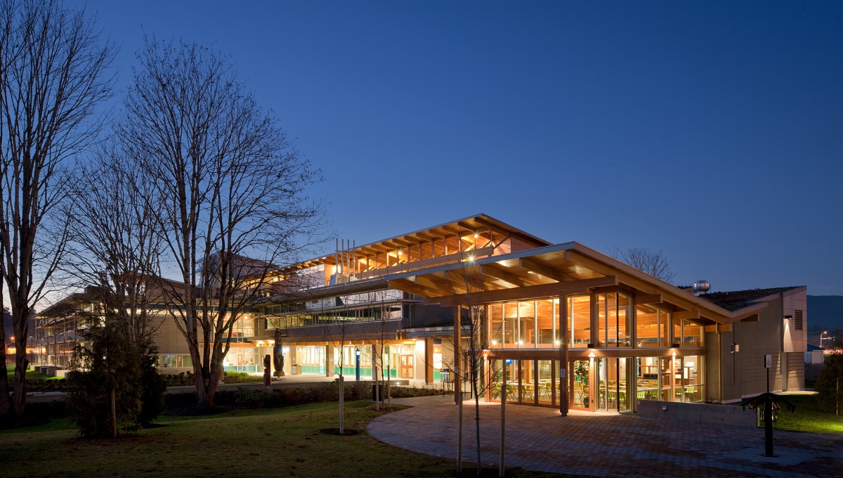 Exterior building wide nighttime view of Vancouver Island University Cowichan Campus showing lit glass building front and including Glue-laminated timber (Glulam) beams and columns supporting expansive wooden roof