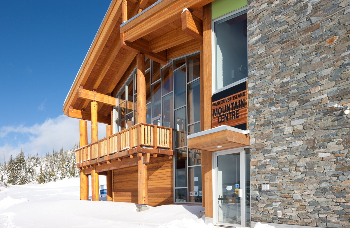 Extensive use of mass timber, including glue-laminated timber (Glulam) beams, wood siding & soffit, solid-sawn heavy timber vertical pole columns, dimensional lumber banister & railing are all shown in this sunny close up outdoor daytime image of the Vancouver Island Mountain Centre building