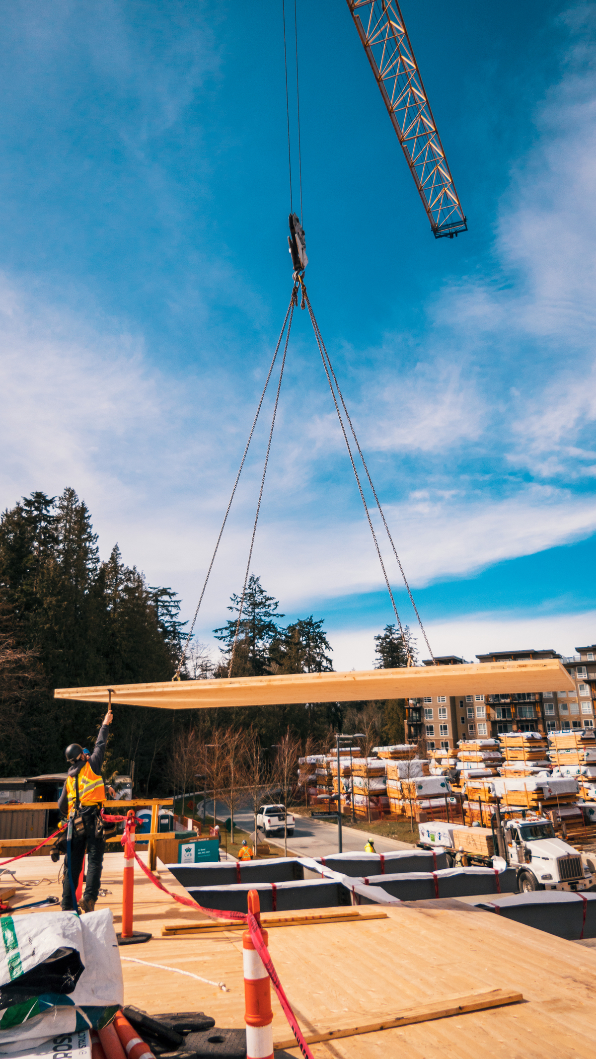 Outdoor daytime image of hybrid multi family residential Virtuoso building during early construction showing cross-laminated timber (CLT) floor panel being lifted into place onto steel beams by crane, with worker in bright PPE guiding