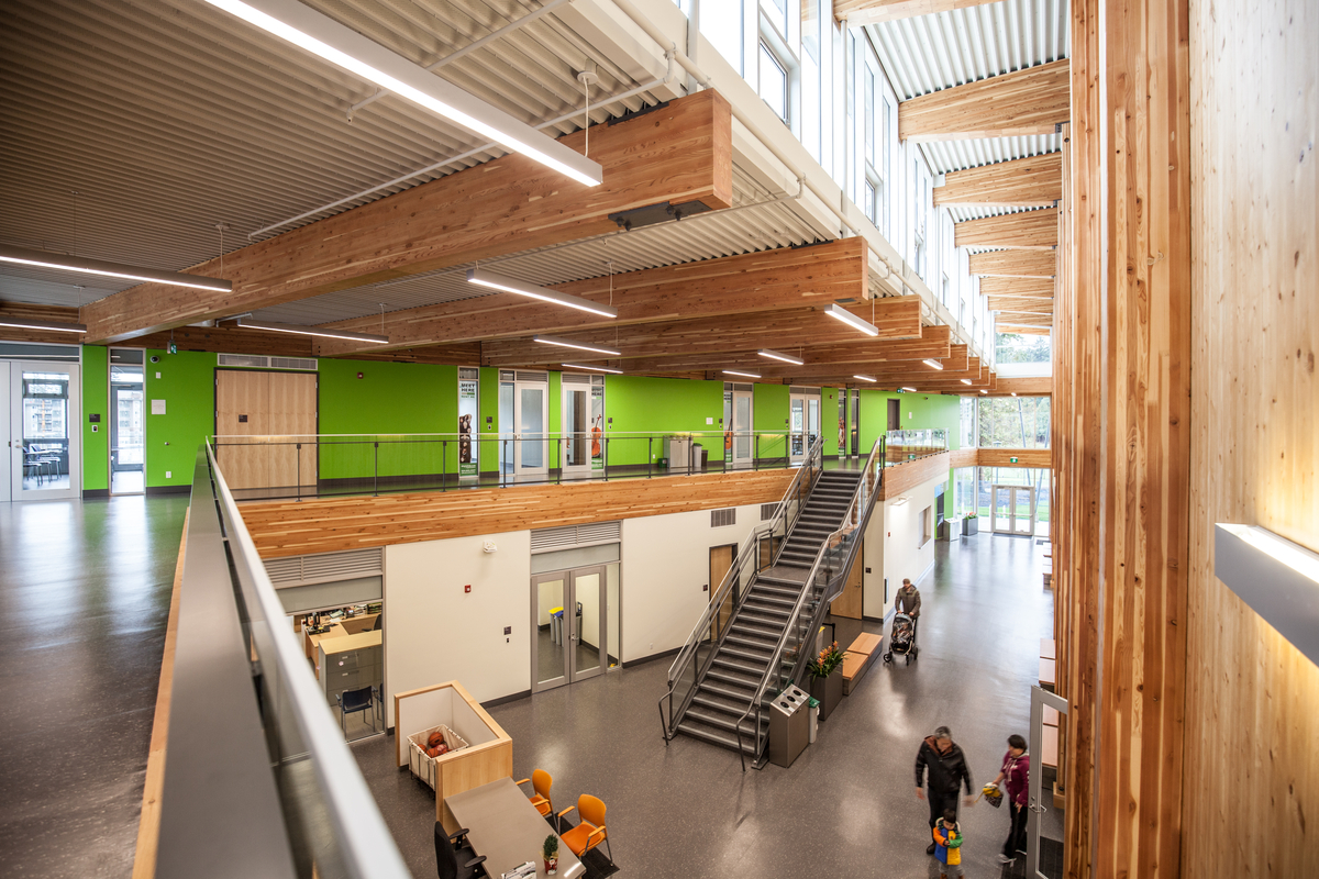 Interior daytime top floor balcony image of occupied multi storey low rise Wesbrook Community Centre main atrium showing cross-laminated timber (CLT) wall paneling and full span horizontal glue-laminated timber (Glulam) ceiling beams, along with extensive use of wood trim