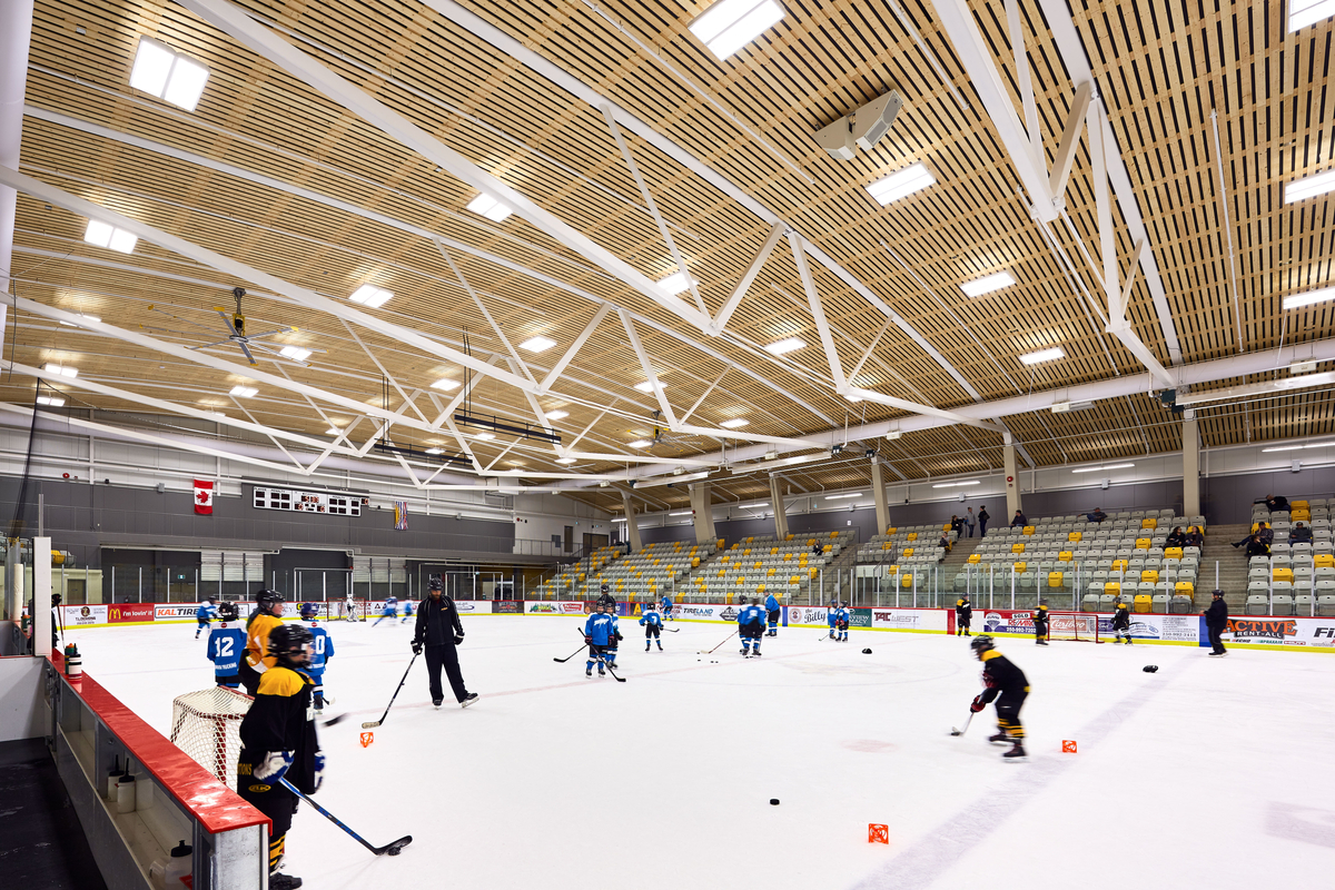 Interior view of West Fraser Centre Ice Rink showing hockey practice and highlighting the large roof structure of the main arena, framed with curved steel girder trusses accented by a wood slat ceiling assembly
