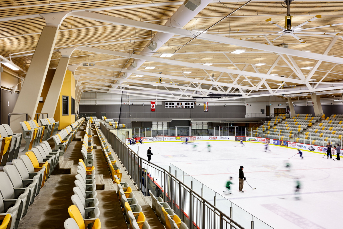 Interior upper seating view of West Fraser Centre Ice Rink showing hockey practice and highlighting the large roof structure of the main arena, framed with curved steel girder trusses accented by a wood slat ceiling assembly