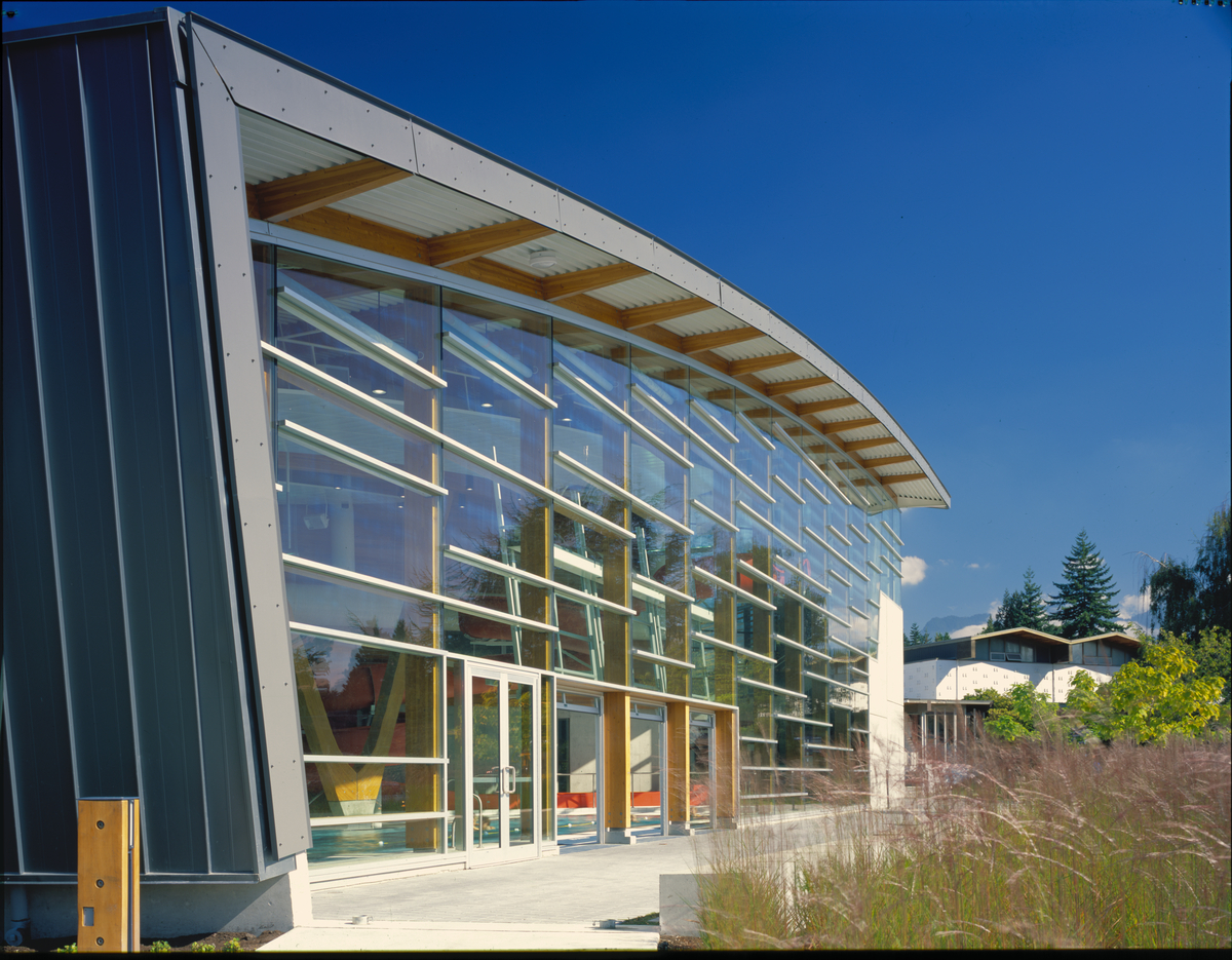 Exterior daytime view of low rise West Vancouver Aquatic Centre looking through floor to ceiling glazed wall panels inward at vertical glue-laminated timber (Glulam) columns supporting glue-laminated timber (Glulam) roof beams which extend to external eaves and soffits