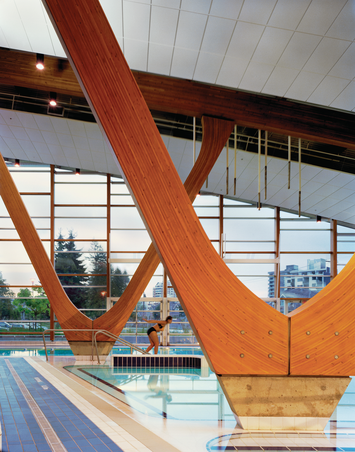 Interior daytime view of low rise West Vancouver Aquatic Centre showing pool surface, swimmer, and curved Glue-laminated timber (Glulam) columns arcing up to support Glue-laminated timber (Glulam) roof beams
