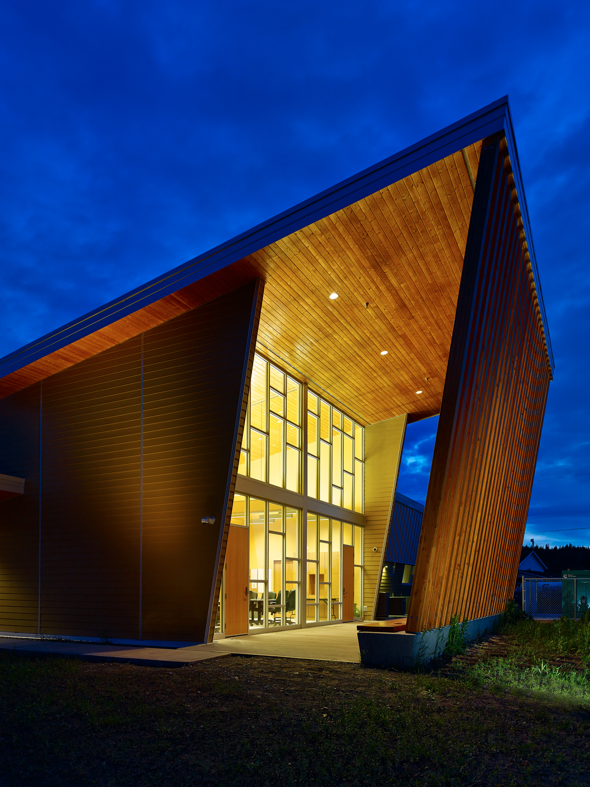 Exterior evening view of YunesitIn Health Centre entrance showing expansive wood and glass use, including plank siding and Glue-laminated timber (Glulam) columns supporting the prefabricated wood roof trusses