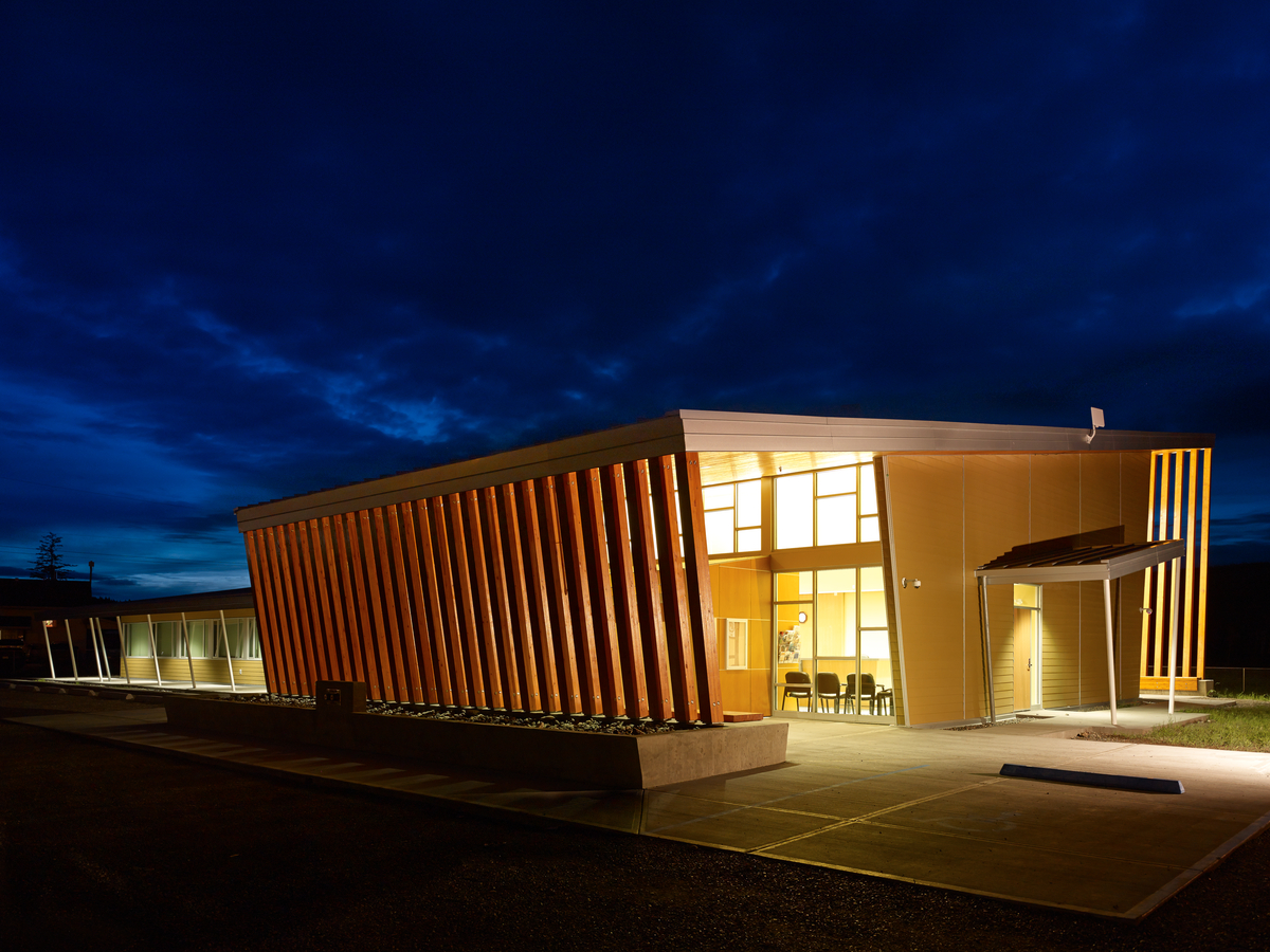 glue-laminated timber (Glulam), Lumber, Millwork, and wooden Siding are all shown in this late evening exterior image of the Yunesit'in Health Centre building and brightly lit main entrance