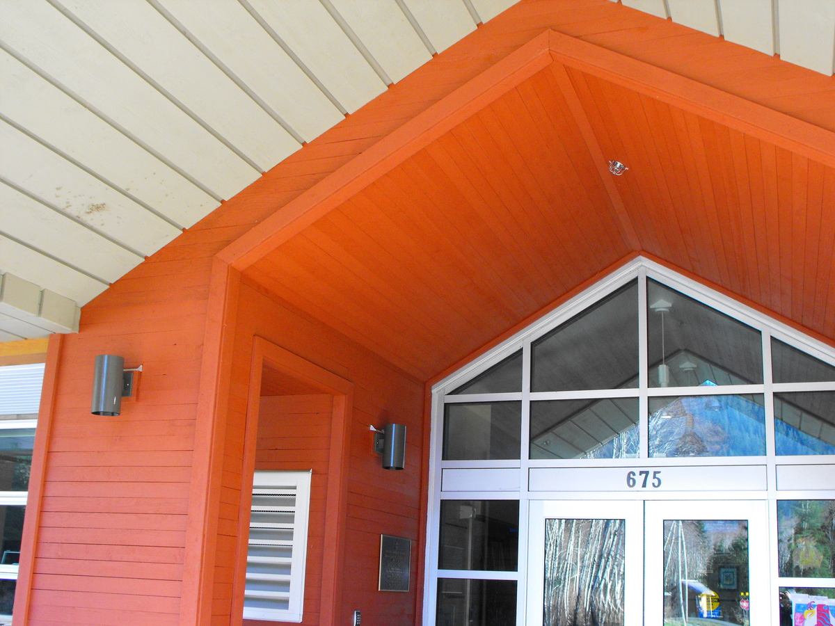 Exterior wood plank paneling, including bright orange accents, are shown in this exterior daytime image of Zeballos Elementary/Secondary School main entrance