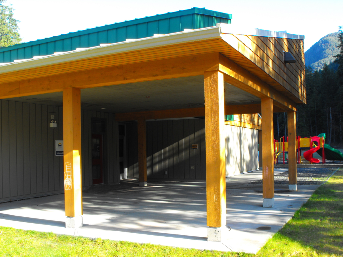 External daytime image of Zeballos Elementary/Secondary School showing wooden covered entrance area with solid-sawn heavy timber columns, lumber ceiling, and wood roof shingles