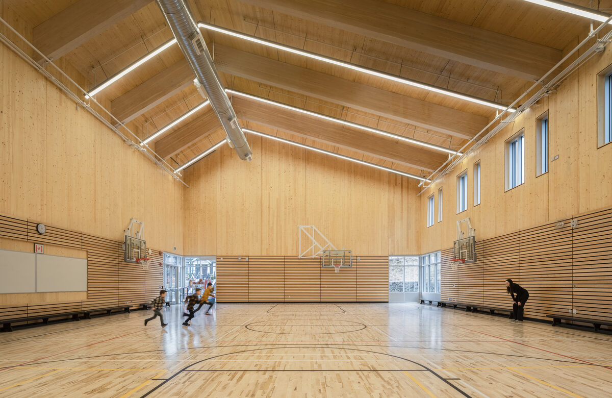 Kids running around in an empty gym. The gym ceilings and walls are all made of wood.