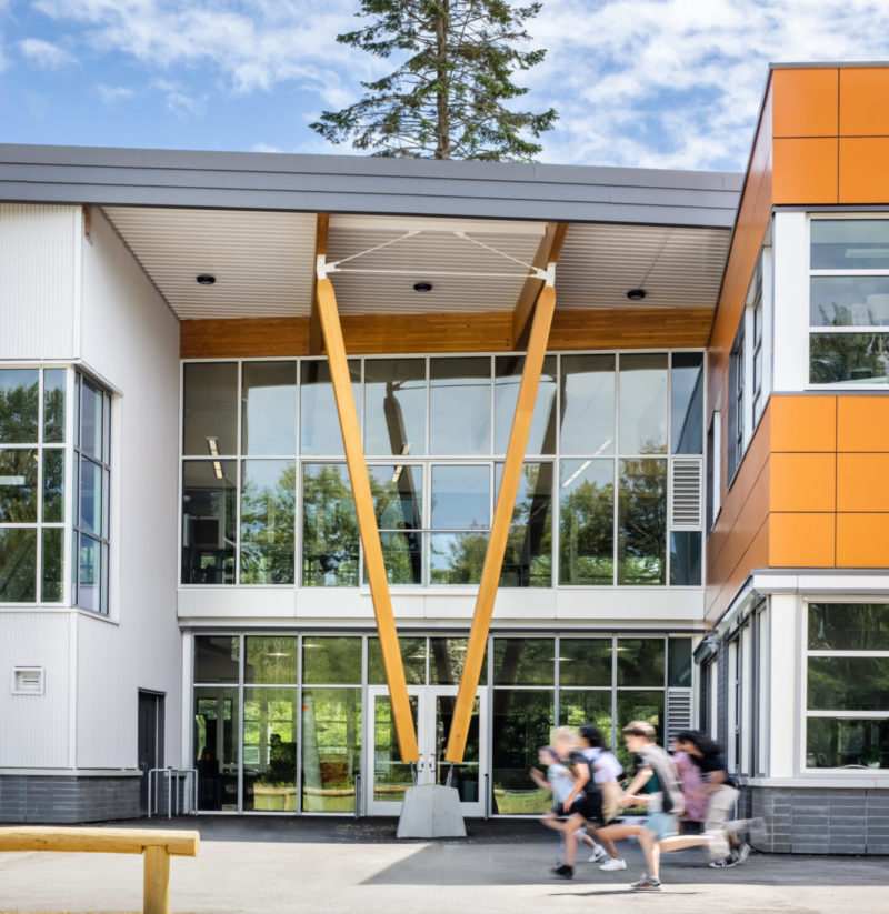 Exterior view of school facade with glass windows covering both entrance floors and V shaped mass timber columns supporting the exterior roof at entrance.