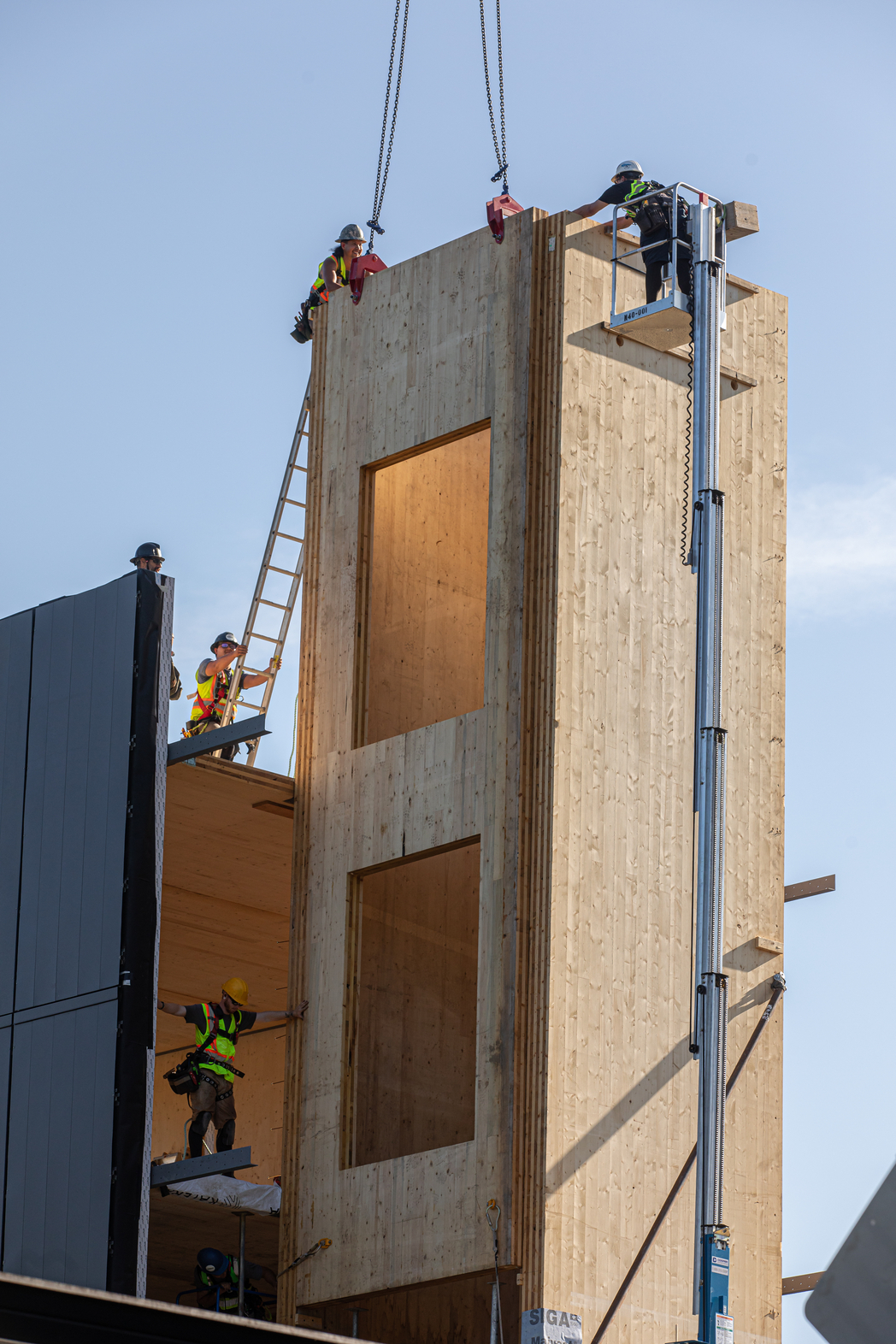 Construction workers align a prefabricated wood panel for elevator shaft while it hangs from a crane.