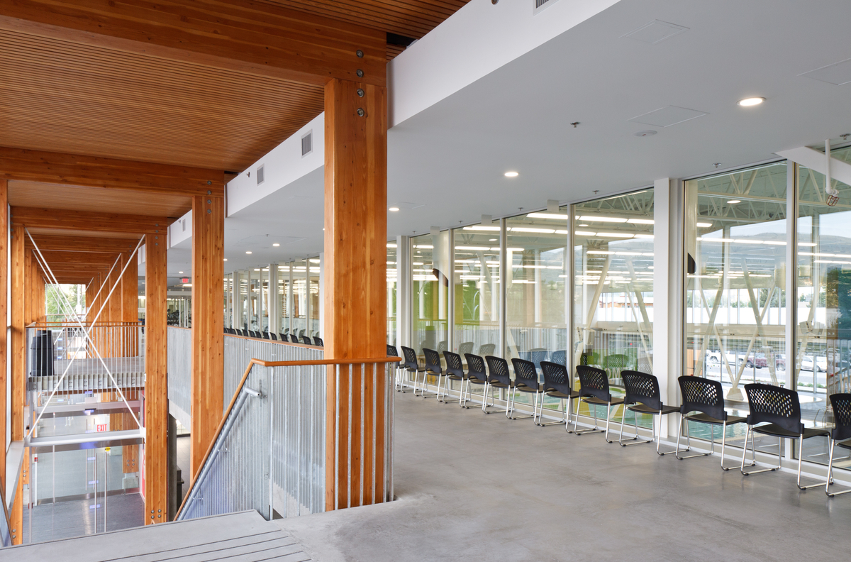 Interior view of Penticton Community Centre top floor with glass separated observation area overlooking pool, including main stairwell with glue-laminated timber (Glulam) columns and slatted Lumber ceiling above