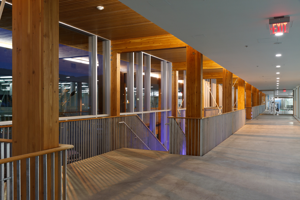 Interior view of Penticton Community Centre top floor showing main stairwell with glue-laminated timber (Glulam) columns and slatted Lumber ceiling above, weight room in background