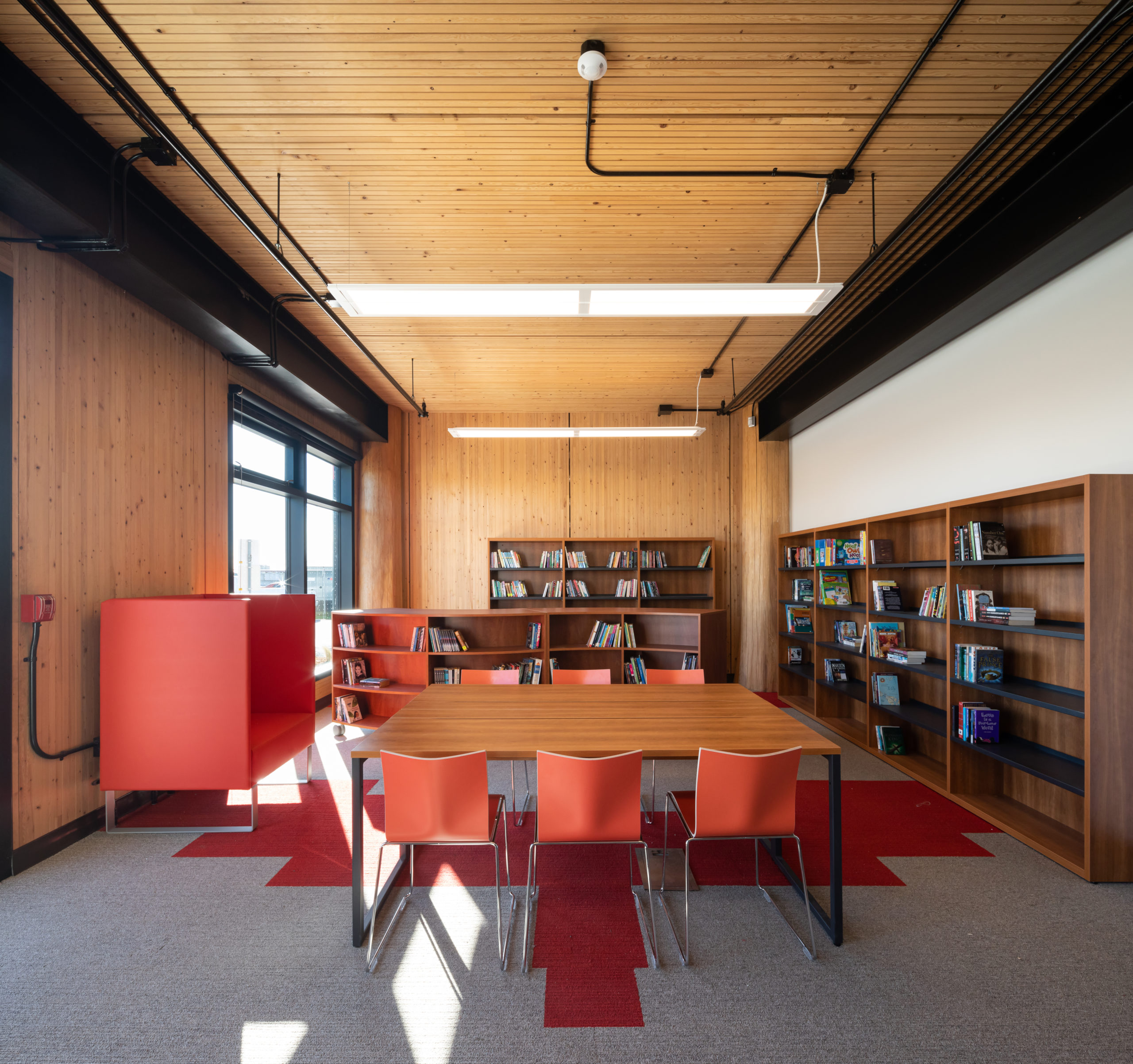 Interior view of library room with exposed wood walls, ceiling, wood book shelves against two walls opposite to a big window. Red accent chairs match the red shapes on the carpet.