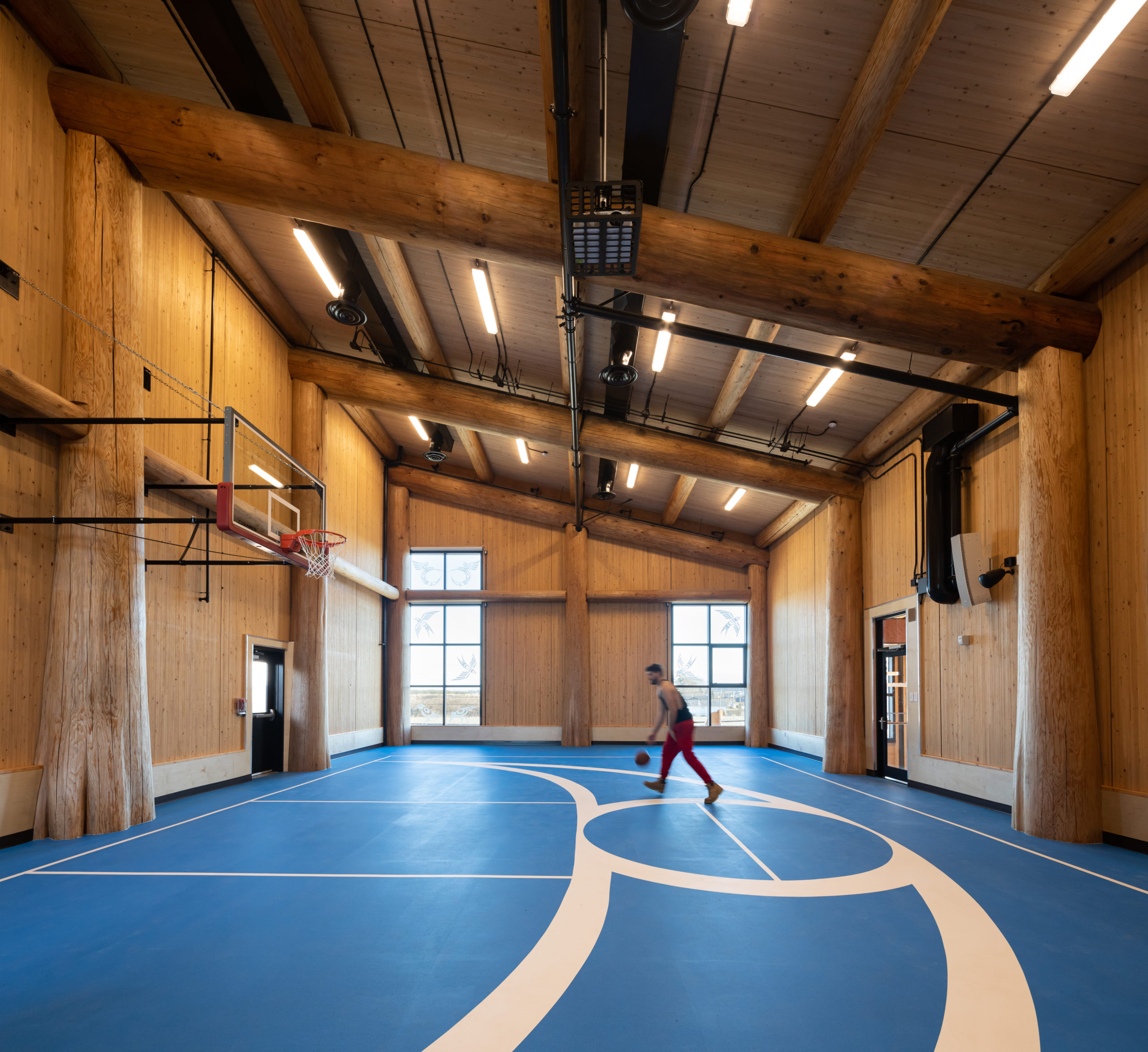 Interior view of gymnasium with slanted roof. The entire walls and ceiling have exposed wood, with large mass timber columns. A young person is playing basketball in the middle of the room with blue flooring with white court marks.